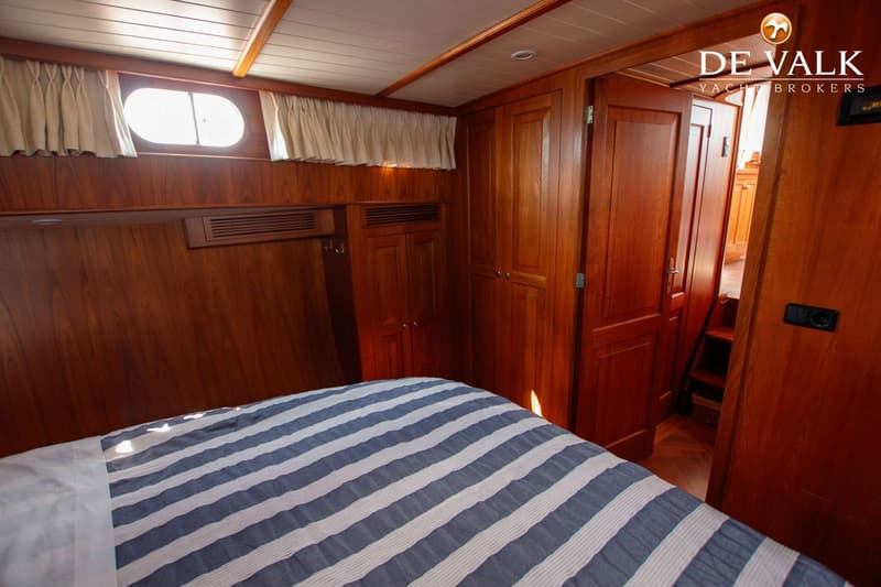 Een kajuit met een gestreept dekbed, houten wanden en kasten, een patrijspoort met gordijnen en een open deur naar een trap.