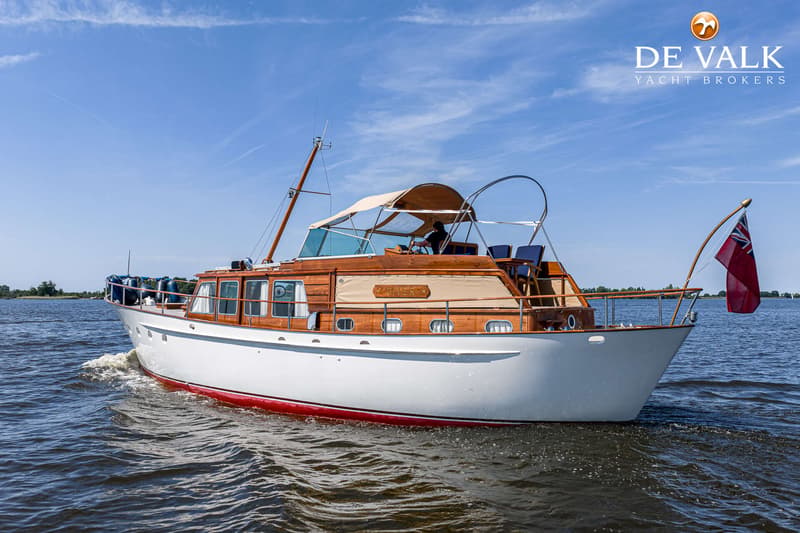 Een motorjacht met een witte romp, rode onderkant en houten opbouw vaart op een waterweg onder een blauwe lucht.