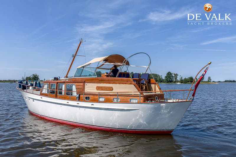 Een witte motorboot met houten opbouw en een beige bimini vaart op een meer onder een blauwe lucht.