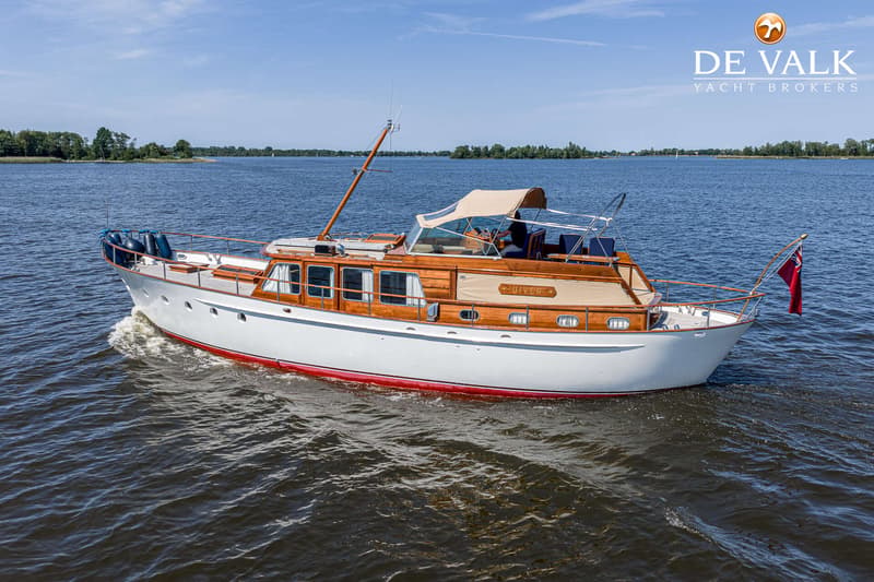 Een witte motorboot met houten opbouw en een rode streep op de romp vaart op een meer onder een blauwe lucht.