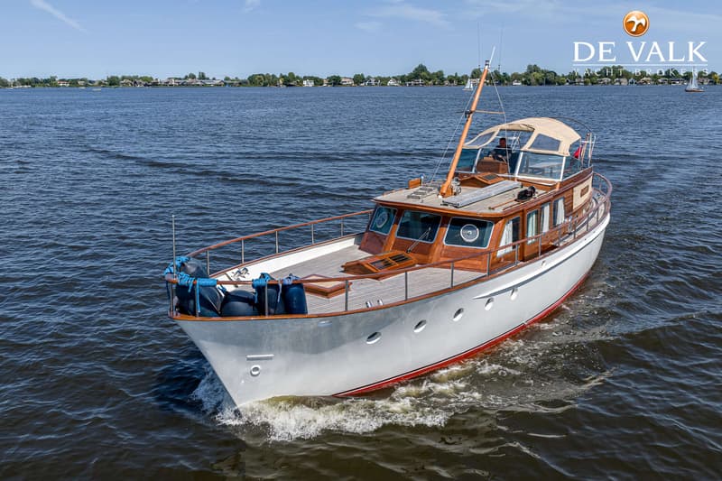 Een witte motorboot met houten opbouw vaart op een meer met huizen aan de oever onder een blauwe lucht.