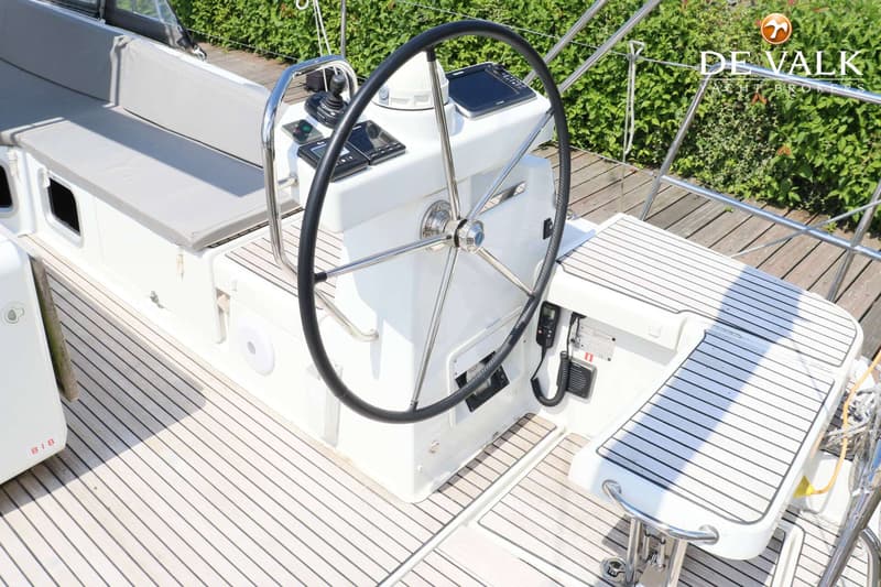Een stuurwiel met instrumentenpanelen, een bank met grijze kussens en een houten dek zijn zichtbaar.