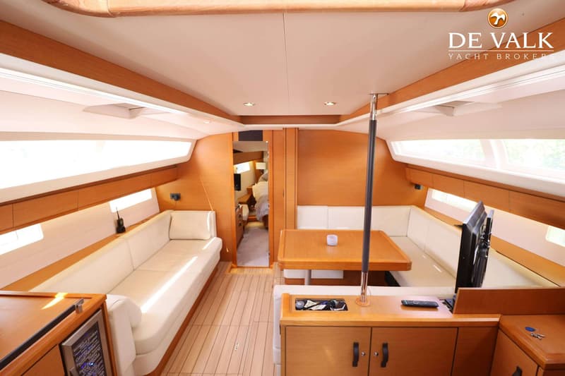De foto toont het interieur van een boot met een houten vloer, witte banken, een houten tafel en een televisie. — Jeanneau 54