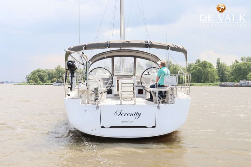 Een witte zeilboot met de naam "Serenity" en "Enkhuizen" op de spiegel, vaart op een rivier met een man aan het roer.