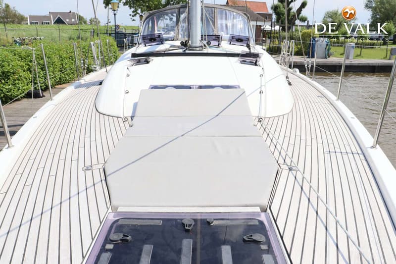 De foto toont het voordek van een zeilboot met een houten dek, een grijze kussenset en een luik. — Jeanneau 54