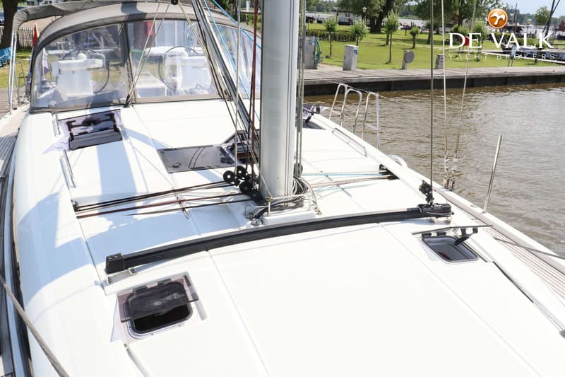 Een wit zeiljacht met een mast, touwen, luiken en een buiskap, liggend aan een steiger met water en groen aan de overkant.