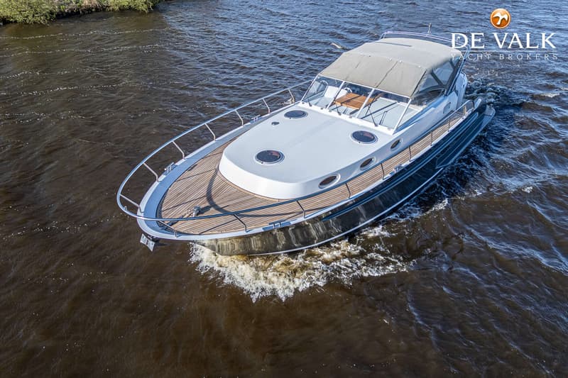 Een motorjacht met een donkere romp en een licht dek vaart op donker water, met een logo van "DE VALK YACHT BROKERS"…