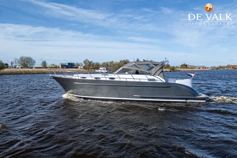 Een grijze motorboot met een wit bovendek vaart op een waterweg onder een blauwe lucht met wolken.