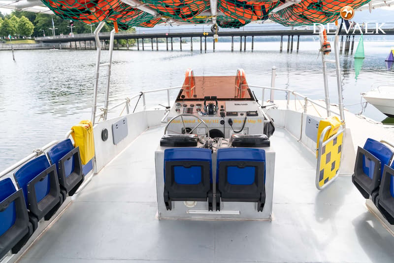 Een bootdek met stuurstand, blauwe stoelen en reddingsvesten in een net onder het dak, met water en een brug op de…