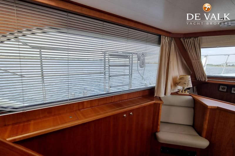 Een interieur van een boot met houten lambrisering, een raam met horizontale jaloezieën en een beige bankje.