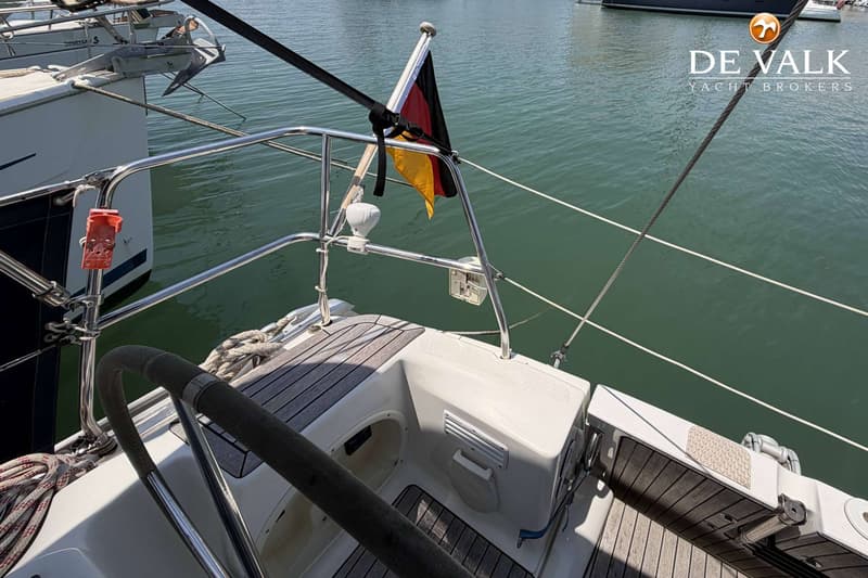 Een deel van een bootdek met een stuurwiel, een Duitse vlag en een reling, met water op de achtergrond.