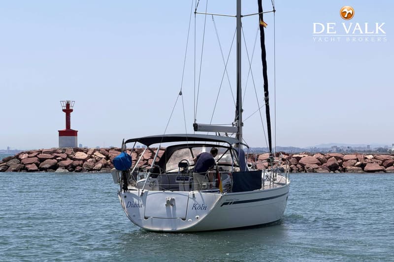 Een witte zeilboot met de namen "Diana" en "Köln" op de spiegel vaart langs een stenen pier met een rood-witte vuurtoren.