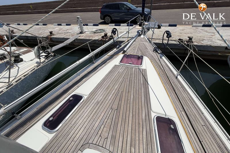 Een bovenaanzicht van het teakhouten dek van een zeilboot, met luiken en een reling, afgemeerd aan een kade.