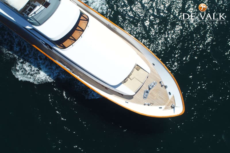 Een bovenaanzicht van een witte motorjacht met een houten dek en oranje accenten, varend op donkerblauw water.