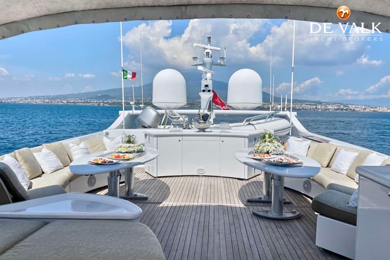 Een achterdek van een jacht met twee tafels, banken, een bar en uitzicht op de zee en een kustlijn.
