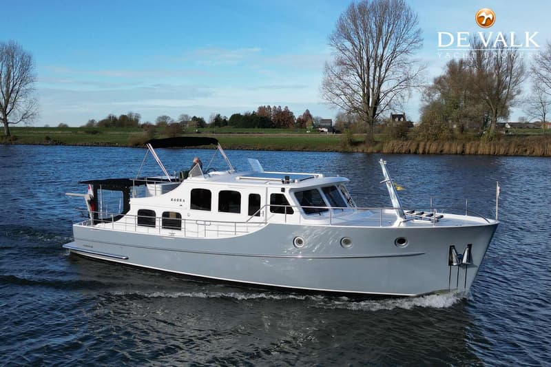 Een grijze en witte motorboot met de naam "KOBRA" vaart over een waterweg met een groene oever en bomen op de achtergrond.