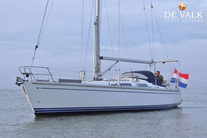 Een witte zeilboot met blauwe strepen op de romp vaart op rustig water, met een man aan het roer en een Nederlandse vlag.