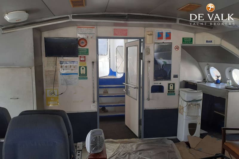 De foto toont het interieur van een boot met zitplaatsen, een bar, een tv-scherm en een trap naar een lager dek.