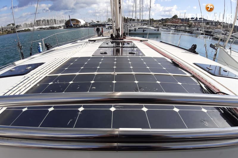 Een dek van een zeilboot met zonnepanelen, luiken, touwen en een mast, met een jachthaven op de achtergrond.
