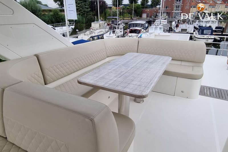 Een bootdek met een U-vormige bank en een houten tafel, met andere boten en gebouwen op de achtergrond.