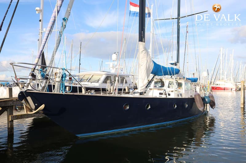 Een donkerblauwe zeilboot met een Nederlandse vlag in een jachthaven, met andere boten op de achtergrond.