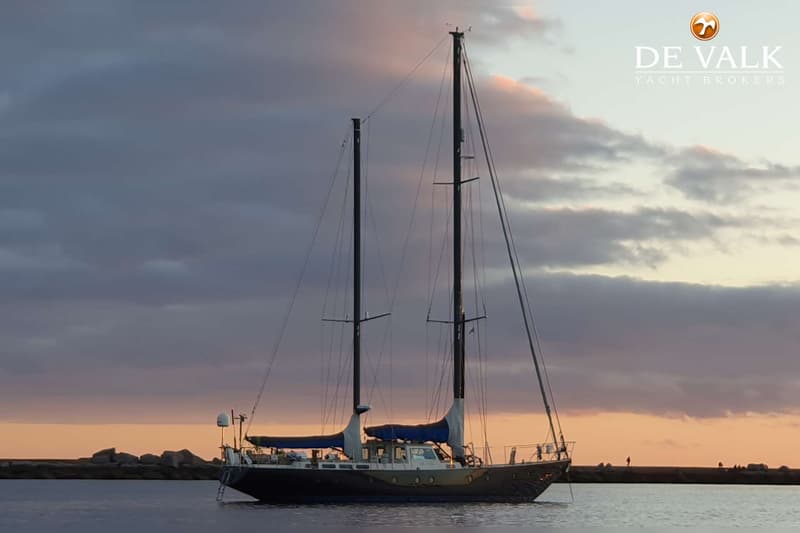 Een donkere zeilboot met twee masten ligt op het water bij zonsondergang, met een rotsachtige kustlijn op de achtergrond.