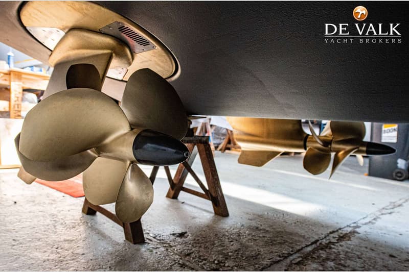 Twee bronzen scheepsschroeven met zwarte naven zijn zichtbaar onder een donkere scheepsromp, ondersteund door houten bokken.