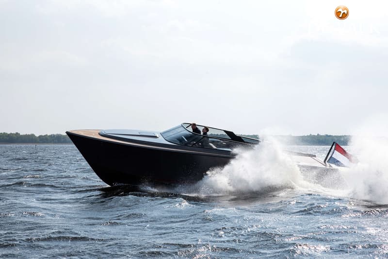 Een donkere speedboot met twee personen aan boord vaart snel over het water, met een Nederlandse vlag aan de achterkant.