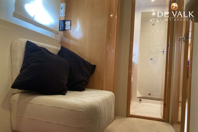 Een interieurfoto toont een beige bankje met twee donkere kussens, naast een houten wand met een open deur naar een…
