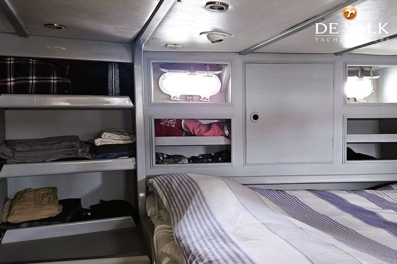 Een kajuit met een bed met gestreepte deken, opbergplanken met kleding en een patrijspoort met licht.