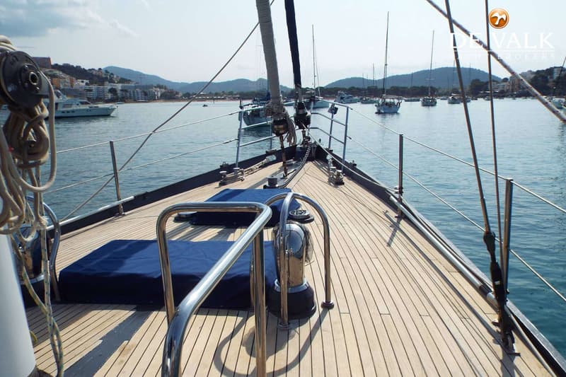 Een houten dek van een zeilboot met blauwe kussens en relingen, met op de achtergrond een baai met andere boten en bebouwing.