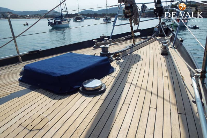 Een teakdek met een blauw afdekzeil, een ankerlier en een bolder, met op de achtergrond een baai vol boten.