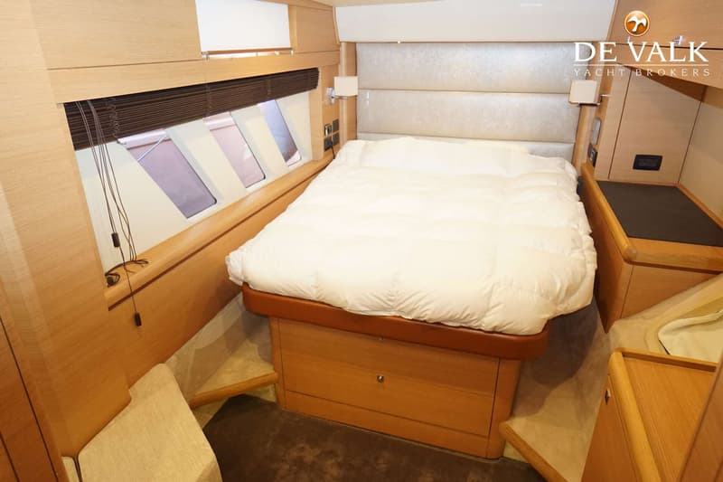 Een slaapkamer in een boot met een groot bed, houten kasten, ramen met jaloezieën en een logo van "DE VALK YACHT BROKERS".