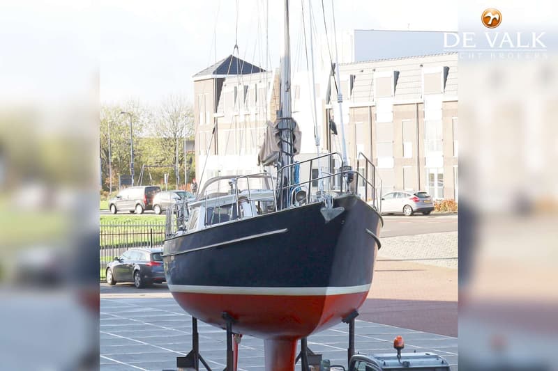 Een donkerblauwe zeilboot met rode onderkant staat op bokken op een parkeerplaats, met gebouwen en auto's op de achtergrond.
