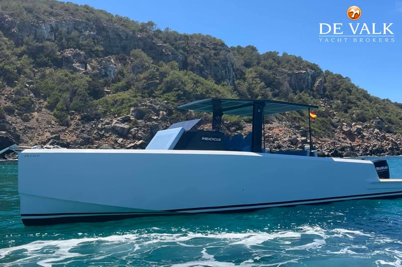 Een witte motorboot met een donkerblauw dak en de tekst "S+BEACHCLUB" op de zijkant, vaart op helderblauw water.