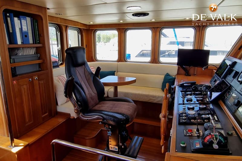 Een interieur van een boot met een stuurstand, een kapiteinsstoel, een bank en een boekenkast.