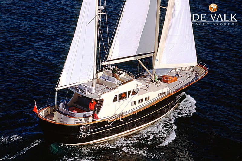 Een donkerblauwe zeilboot met witte zeilen vaart op open water, met een persoon aan het roer en twee personen achterop.