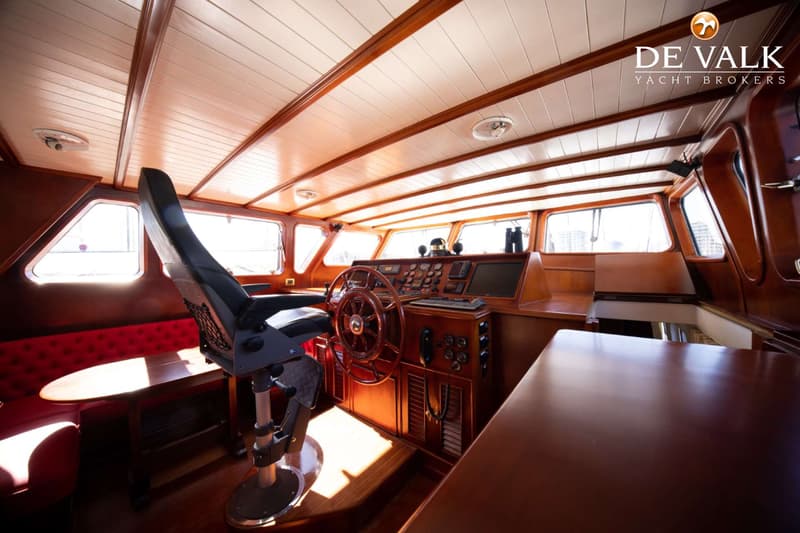 De foto toont een houten stuurhut met een stuurwiel, navigatieapparatuur, een stuurstoel en een rode bank met tafel.