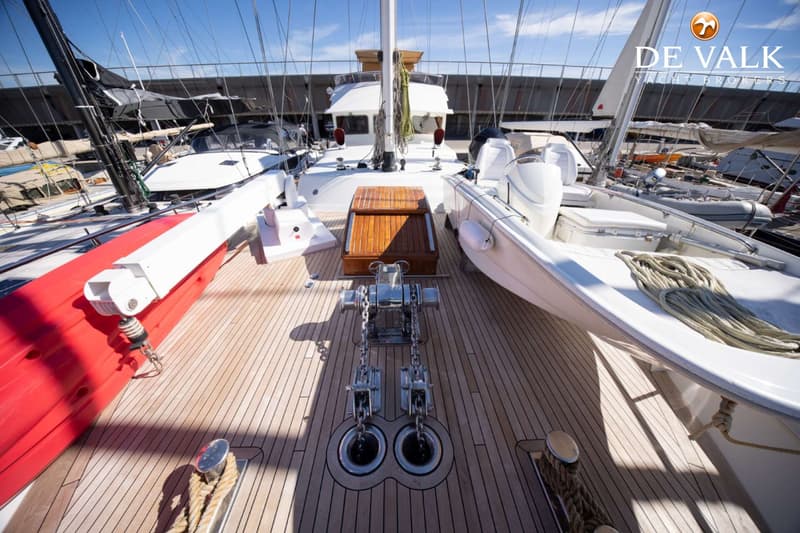 Een bovenaanzicht van het teakhouten dek van een boot met een ankerlier, twee ankerkettingen en een bijboot aan stuurboord.