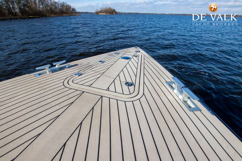 De boeg van een boot met een houten dek en vier bolders, varend op een meer met bomen op de achtergrond. — VanDutch 40