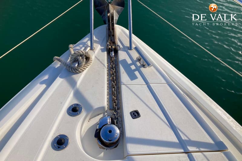 De boeg van een witte boot met een anker, ankerketting, ankerlier en een touw op een bolder, met groen water op de…