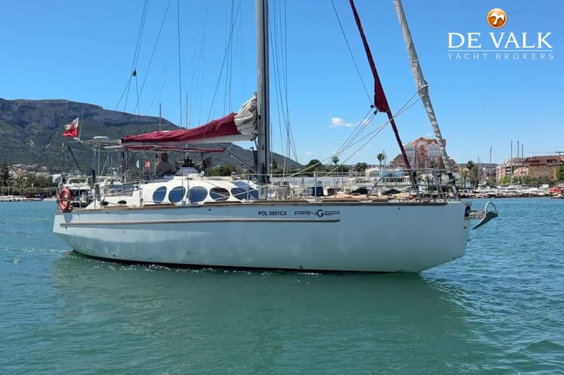 Een witte zeilboot met rode zeilen en de naam "PANGEA" vaart op helderblauw water, met bergen en een stad op de achtergrond.