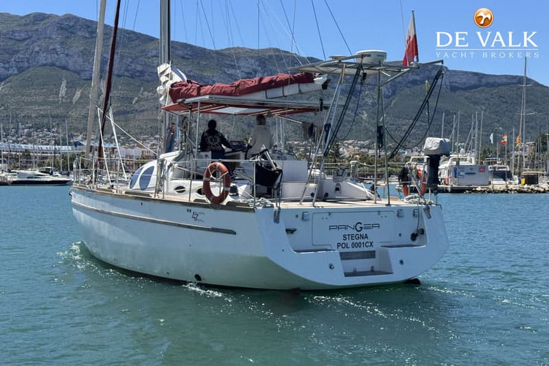 Een witte zeilboot met twee personen aan het roer vaart in een haven met bergen op de achtergrond. — Custom Built Audax 47
