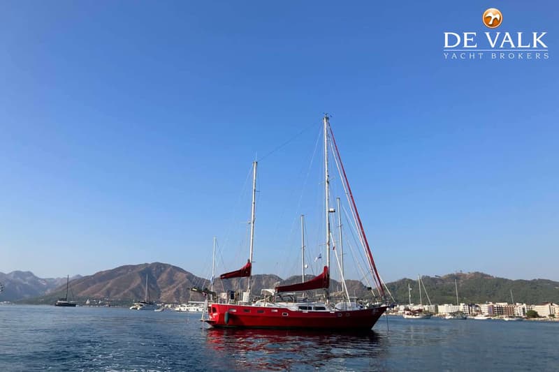 Een rode zeilboot met twee masten en opgerolde rode zeilen ligt in het water, met bergen en bebouwing op de achtergrond.