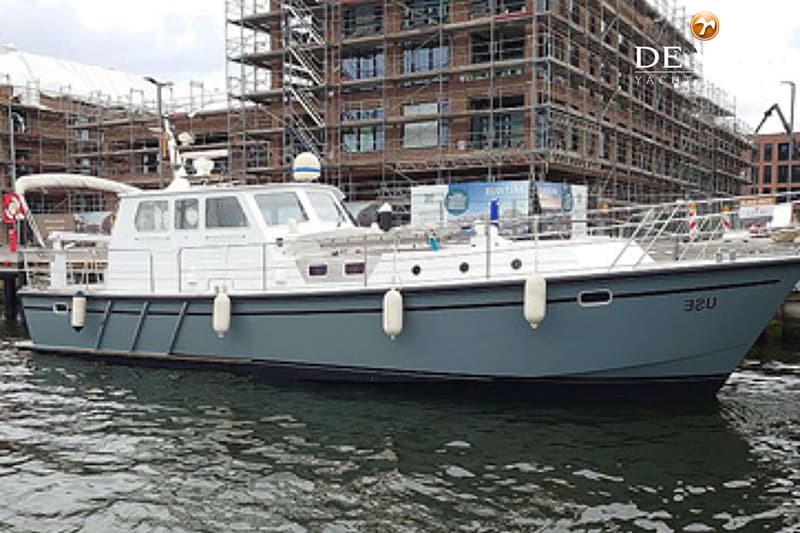 Een grijze motorboot met een witte opbouw en stootwillen drijft in het water voor een gebouw in aanbouw. — Pilot 44