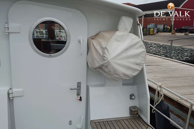 Een witte bootdeur met een rond patrijspoortje en een afgedekt reddingsvlot, naast een houten steiger.