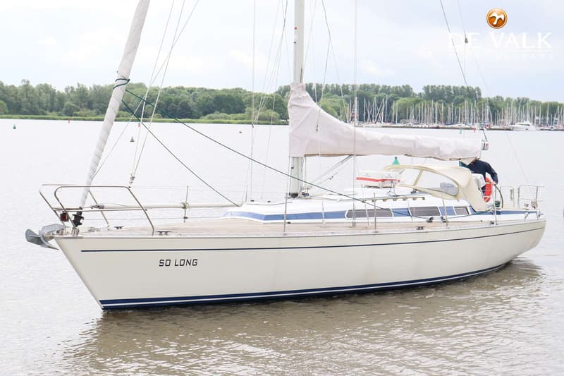 Een witte zeilboot met de naam "SO LONG" vaart op water, met een bosrijke oever en andere boten op de achtergrond.