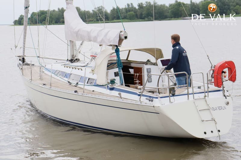 Een witte zeilboot met blauwe strepen vaart op het water, met een persoon aan het roer en een opgerold zeil. — Faurby 396