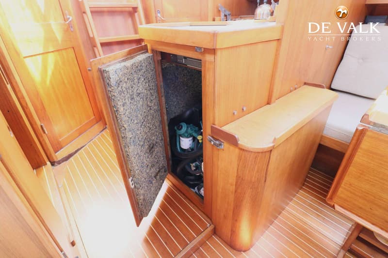 Een houten interieur van een boot met een geopende deur die een motorruimte onthult, en een bank met kussen. — Faurby 396