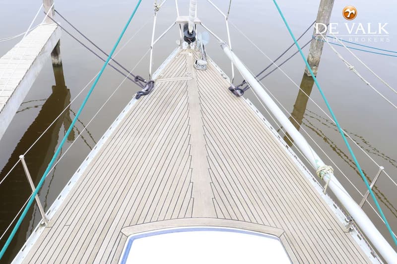 Een bovenaanzicht van het teakhouten dek van een zeilboot met een luik, lijnen en een mast, liggend aan een steiger.
